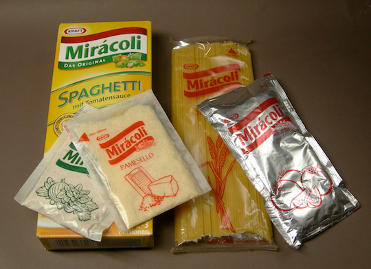 Geöffnetes Päckchen Mirácoli, die vier getrennt abgepackten Bestandteile gut sichtbar nebeneinander gelegt: Gewürzmischung, Hartkäse, Spaghetti, Tomatensauce.