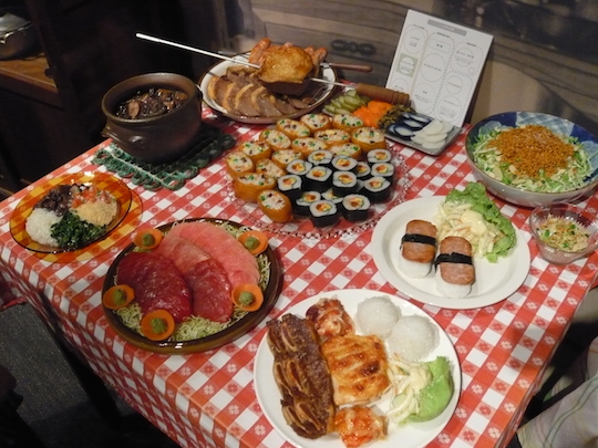 Tisch mit verschiedenen Tellern voller Wachsmodelle von Speisen: Sushi, gebratenes und rohes Fleisch, Salate, Eintopf.