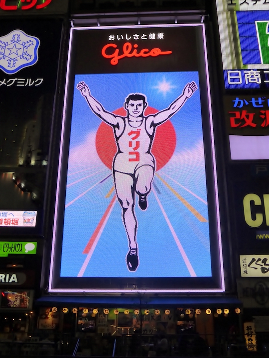 Eine riesige Neonreklame in Ōsaka mit dem so genannten Glico Running Man: oben der Schriftzug oishisa to kenkō („Wohlgeschmack und Gesundheit“) – Glico; darunter der Athlet in weiß vor einem großen roten Punkt im blauen Hintergrund.