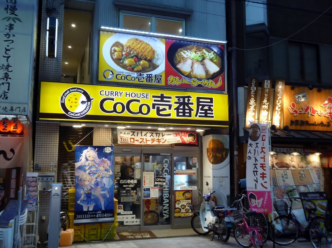 Eine Filiale von Curry Ichiban’ya: schwarzer Schriftzug auf gelbem Grund, darüber große Abbildungen zweier Curry-Gerichte; der Eingang eine gläserne Schiebetür voller Poster und Schilder.