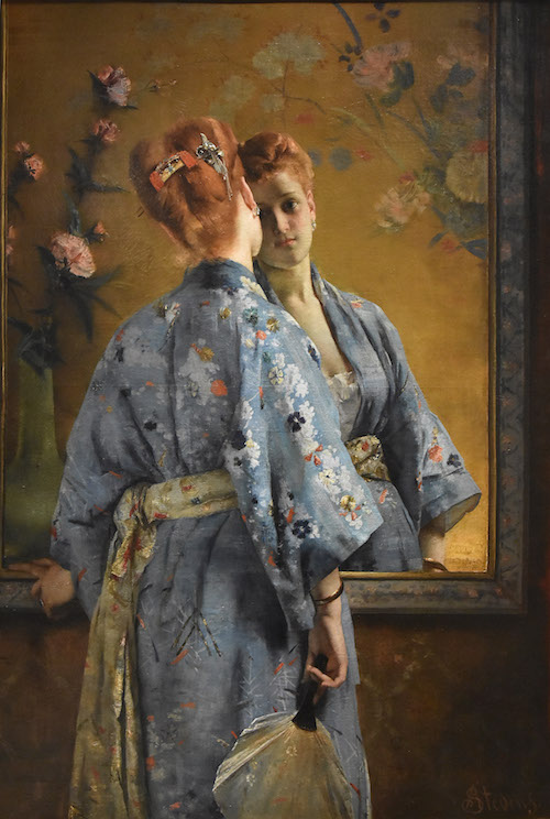 La Parisienne Japonaise von Alfred Stevens ist eine rothaarige Frau, die mit dem Rücken zum Betrachter steht. Sie trägt einen blauen Kimono mit Blumenmuster und hält einen Fächer in der Hand. Im Spiegel sieht der Betrachter ihr Gesicht und ihren weiten Ausschnitt.