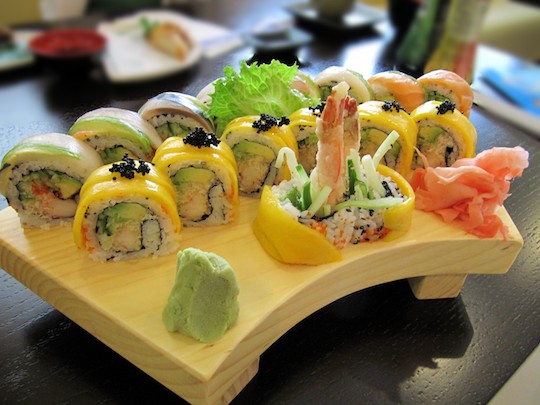 Inside out-Sushi der Rainbow Roll in hellem Grün, Gelb und Zartrosa lassen die Anordnung auf dem Holztablett wie einen Regenbogen erscheinen. Die verwendeten Zutaten sind andere als im Ursprungsland Japan.