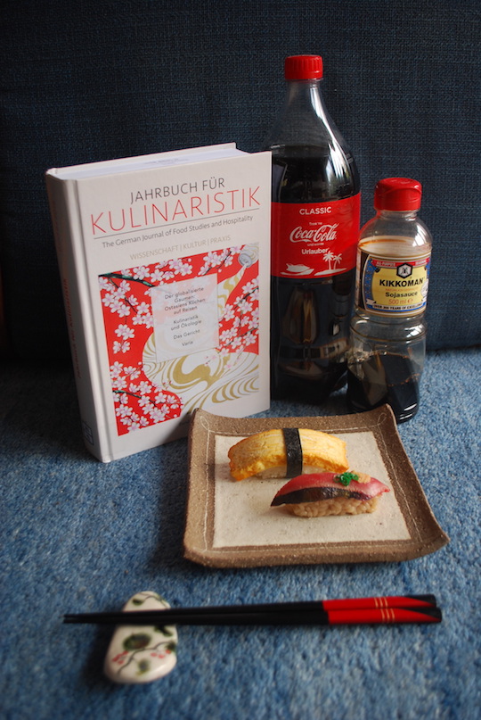 Jahrbuch für Kulinaristik, Cola-Flasche, Flasche Soja-Sauce, Tablett mit zwei Sushi, Stäbchen auf Ablage