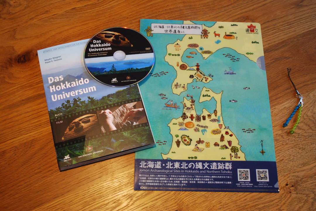 Anordnung von links nach rechts: Das Buch und die DVD mit dem Titel „Das Hokkaidō Universum“; eine Hülle für DinA 4-Papier mit einer handgezeichneten Karte der Grabungsstellen auf Hokkaidō und Nord-Honshū; ein Anhänger mit zwei verdrillten Schnüren in Blau und Grün.
